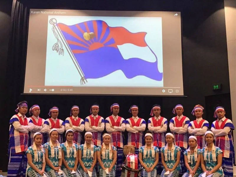 Dancers in traditional Karen dress in two rows on a stage in Melbourne, in front of a screen showing the Karen flag.