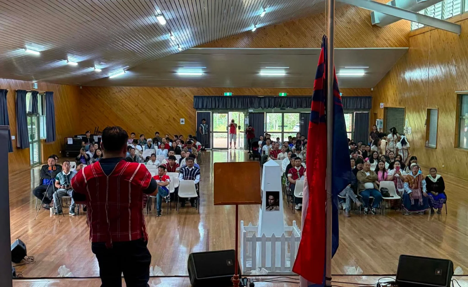A speaker facing a full hall at the Karen Martyrs' Day commemoration, beside the Karen flag and a portrait on a memorial stand.